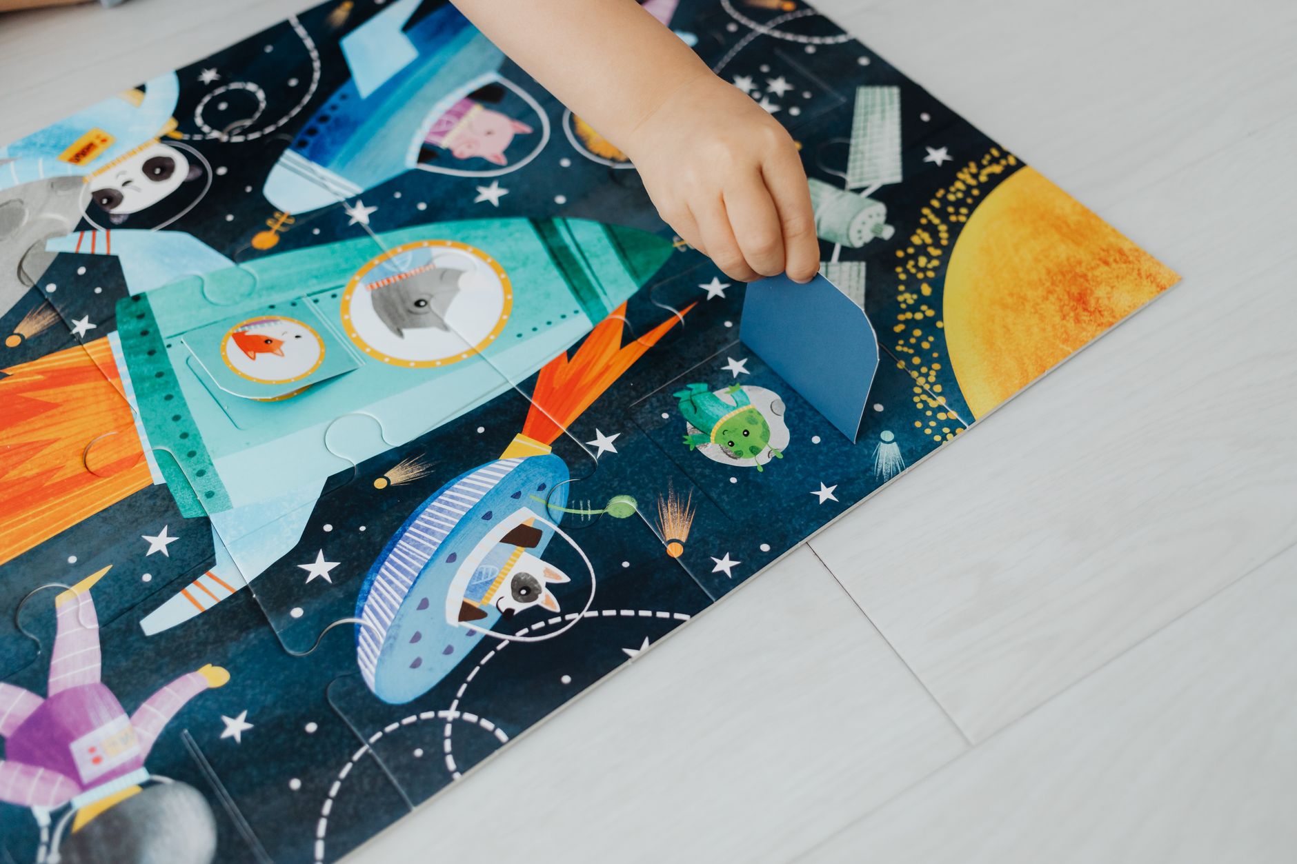 Child's hand assembling a colorful space-themed jigsaw puzzle on a light floor.