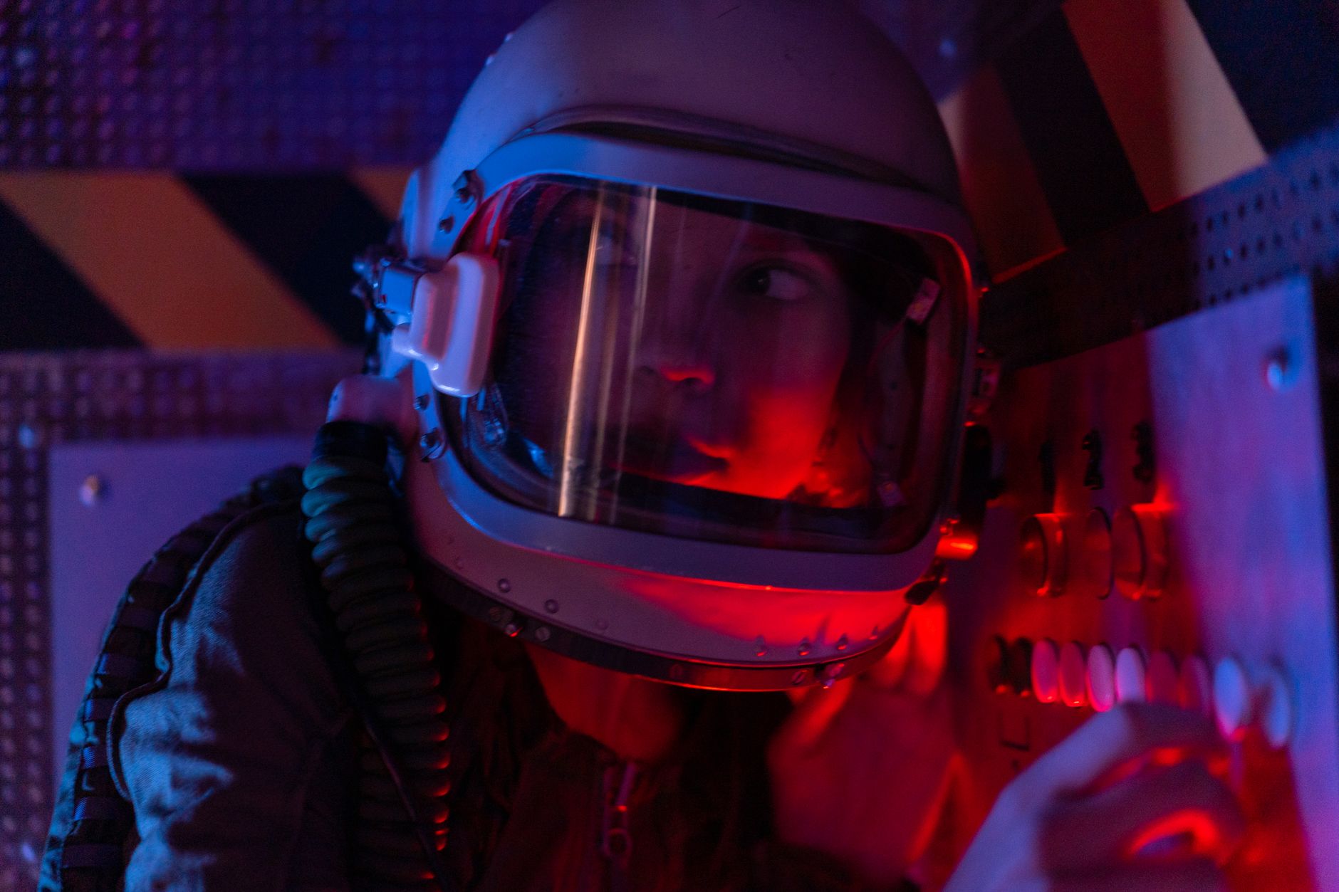 A woman astronaut wearing a spacesuit in a control room lit by red lights, looks focused.