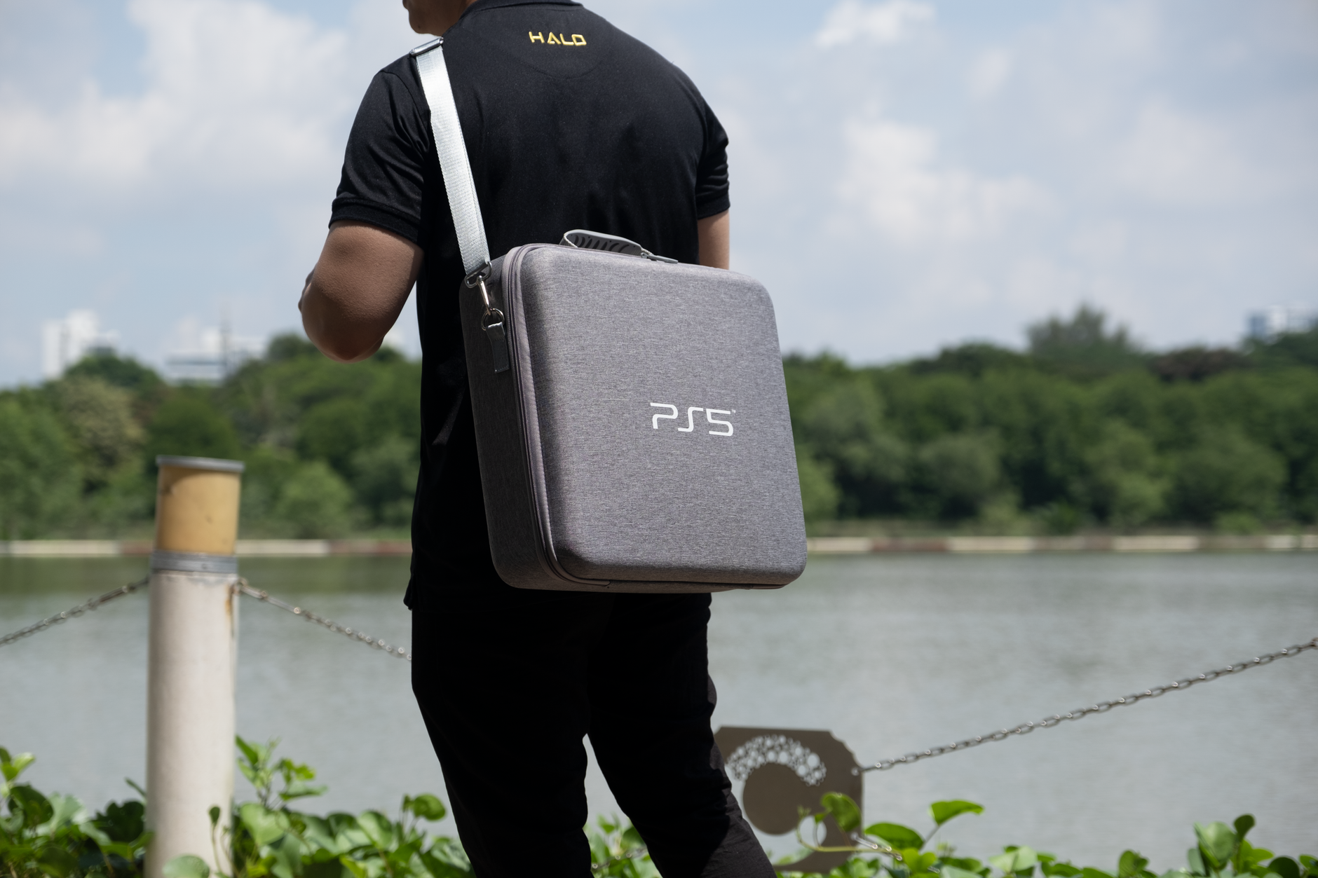 A man carries a PS5 bag by a lake, surrounded by greenery in a park setting.