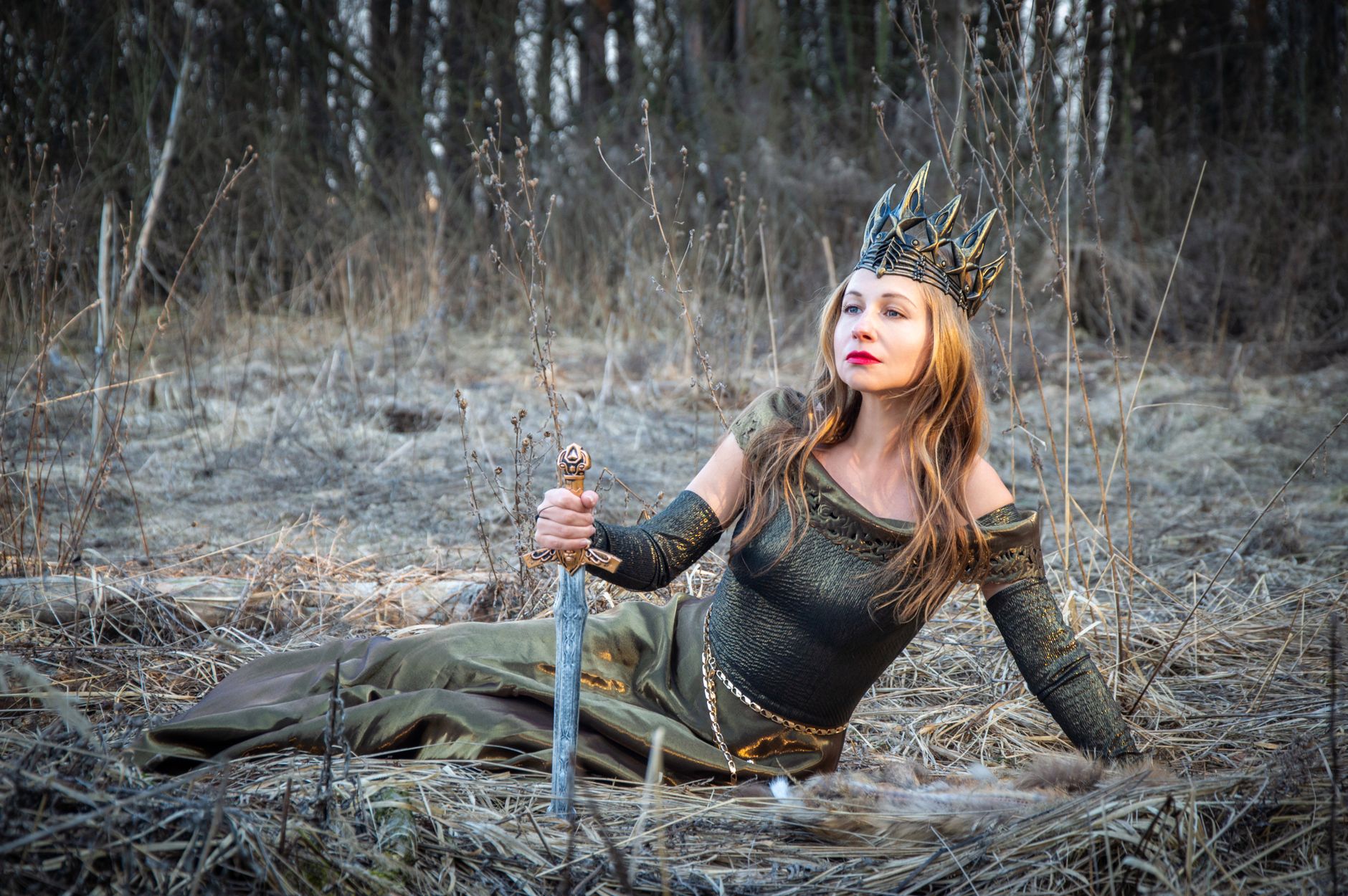A woman dressed as a regal warrior queen with a crown, holding a sword in a mystical forest.