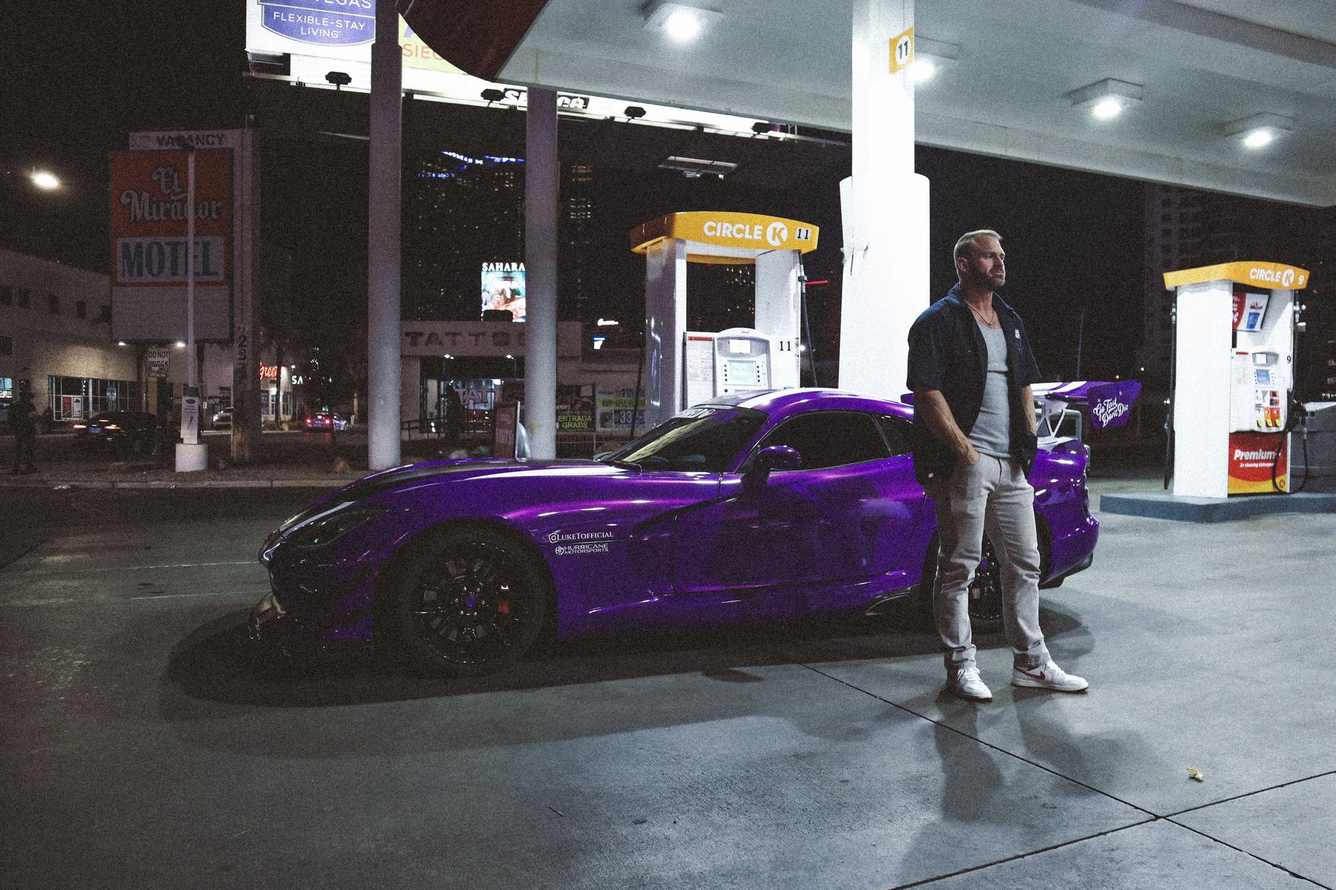 Night scene of a man standing beside a purple sports car at a gas station under city lights.