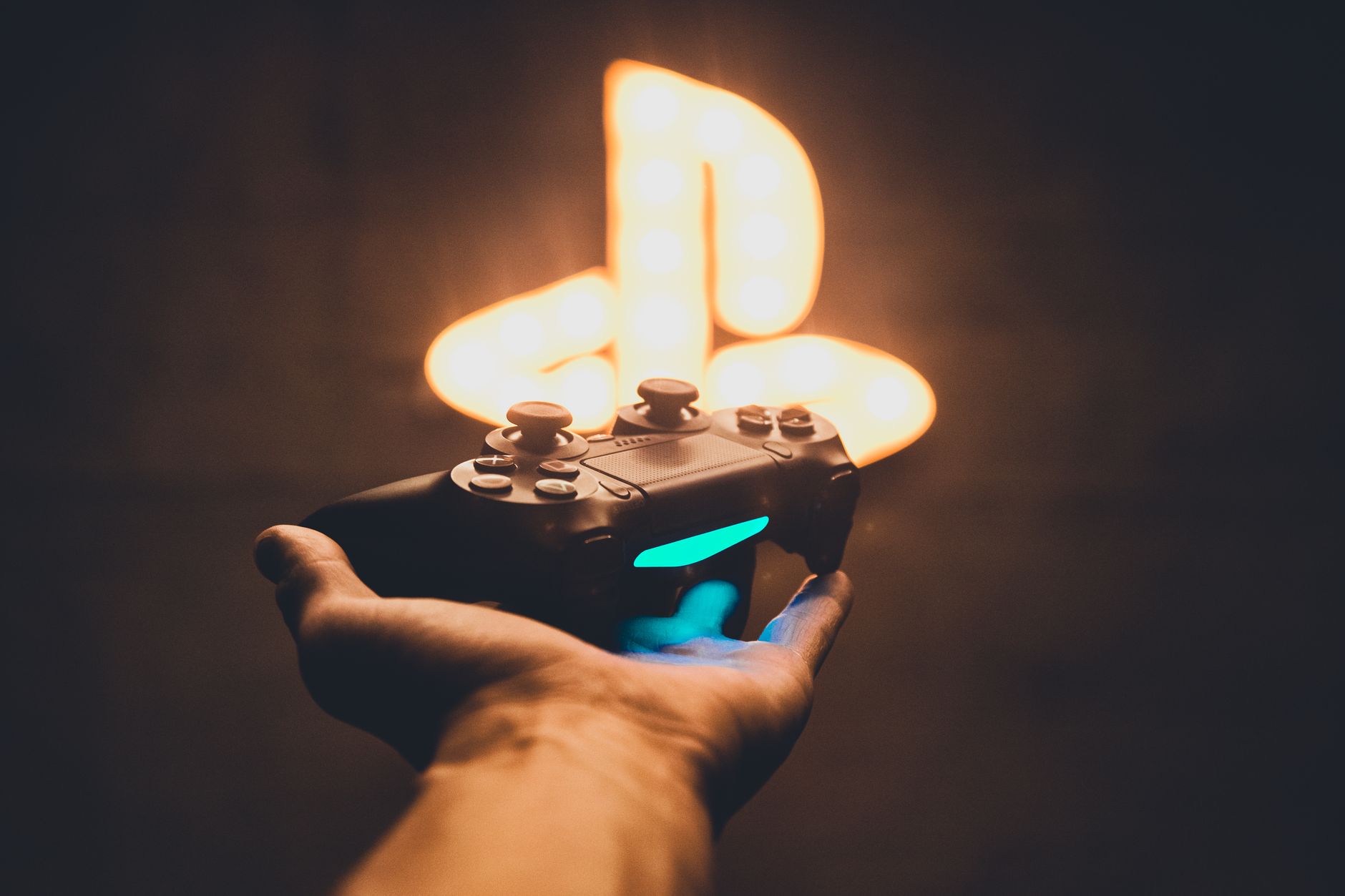 A hand holding a PlayStation controller with a blurred neon game symbol in the background.