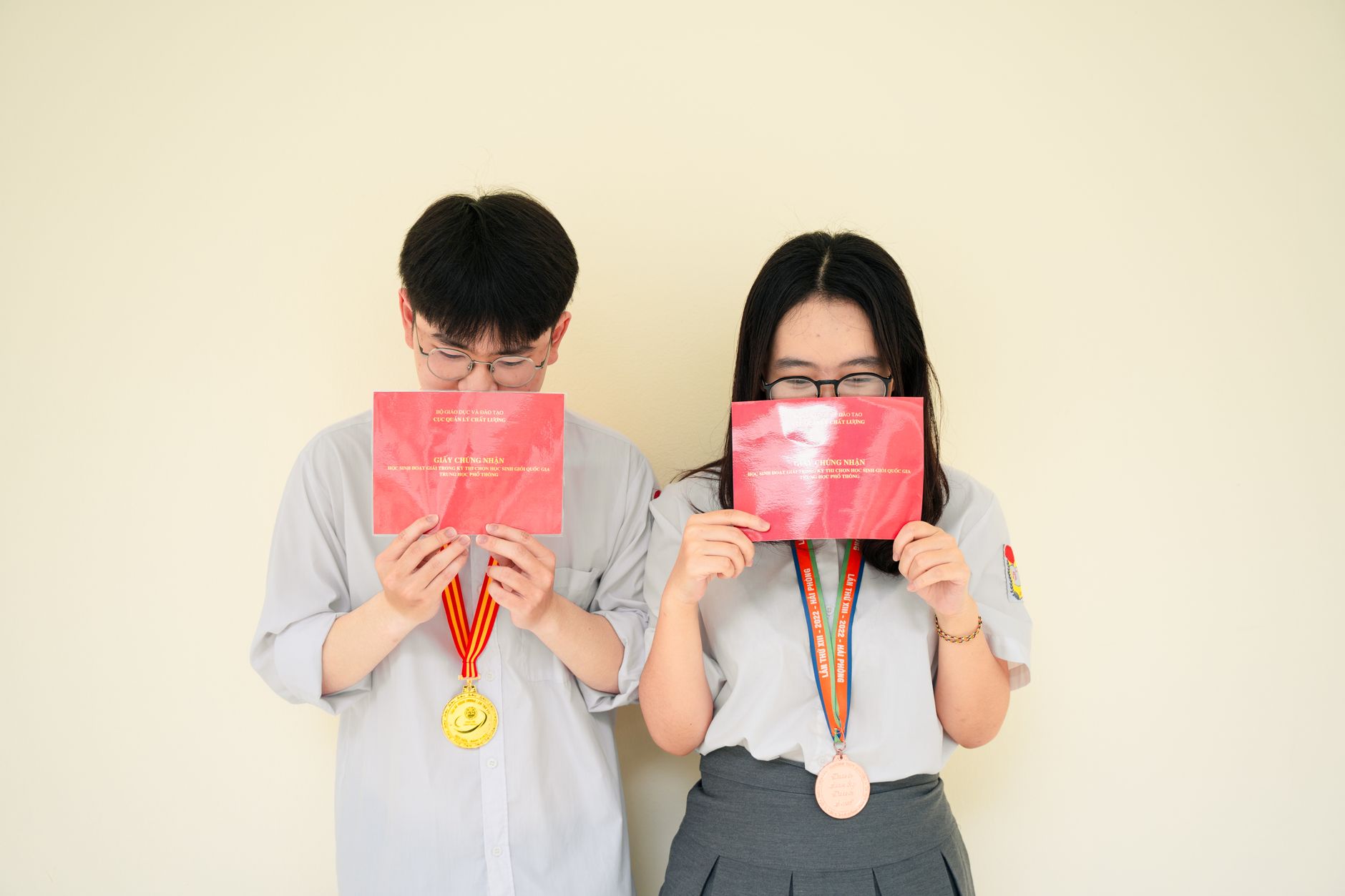 Two Asian teenagers holding diplomas and wearing medals, symbolizing academic success.