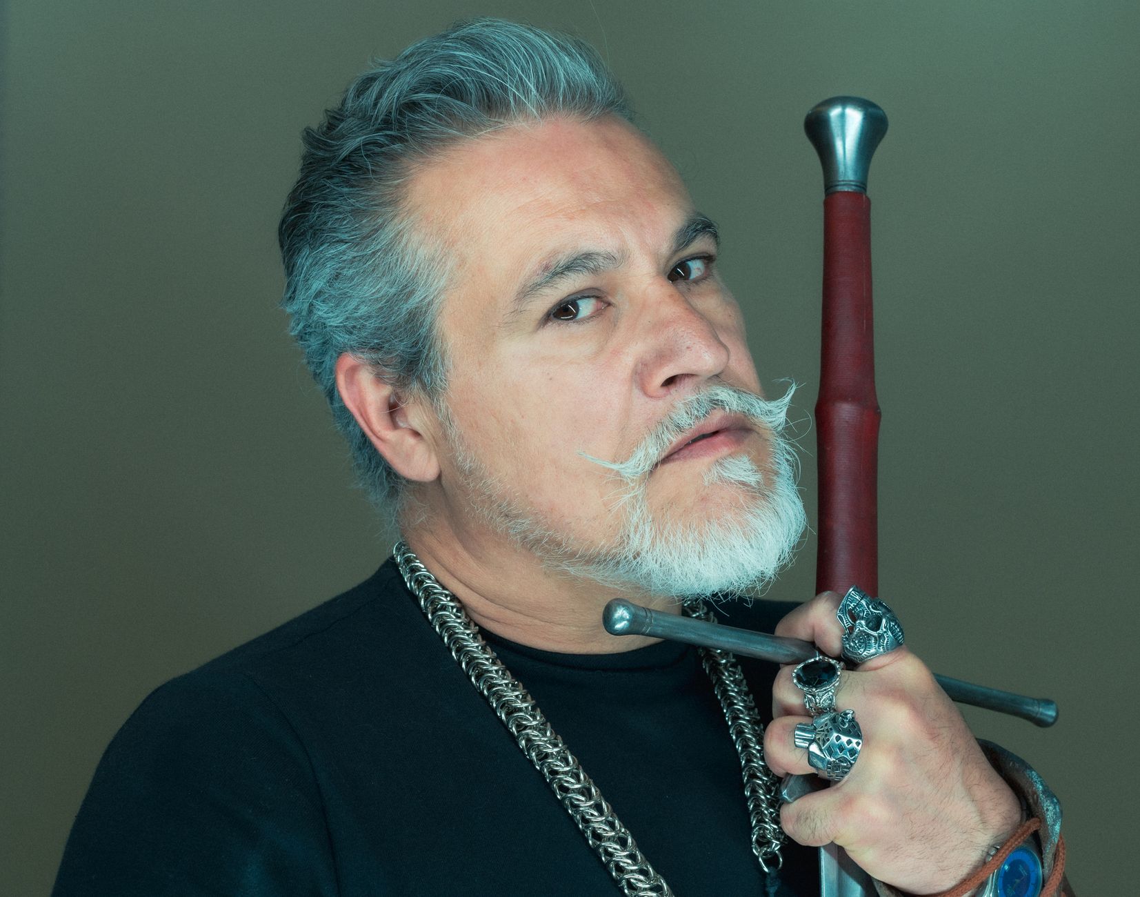 Close-up portrait of a man with a beard and mustache holding a sword, wearing rings.