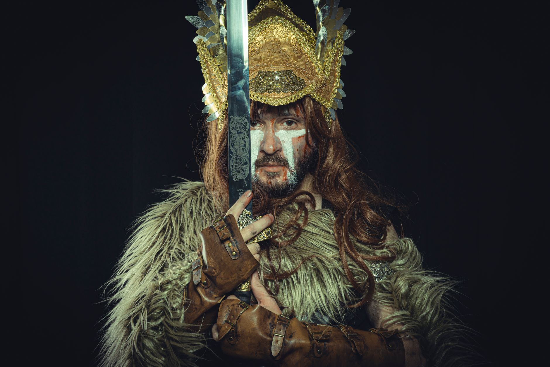 A dramatic portrait of a Viking warrior in costume holding a sword, featuring a golden headdress and fur garment.