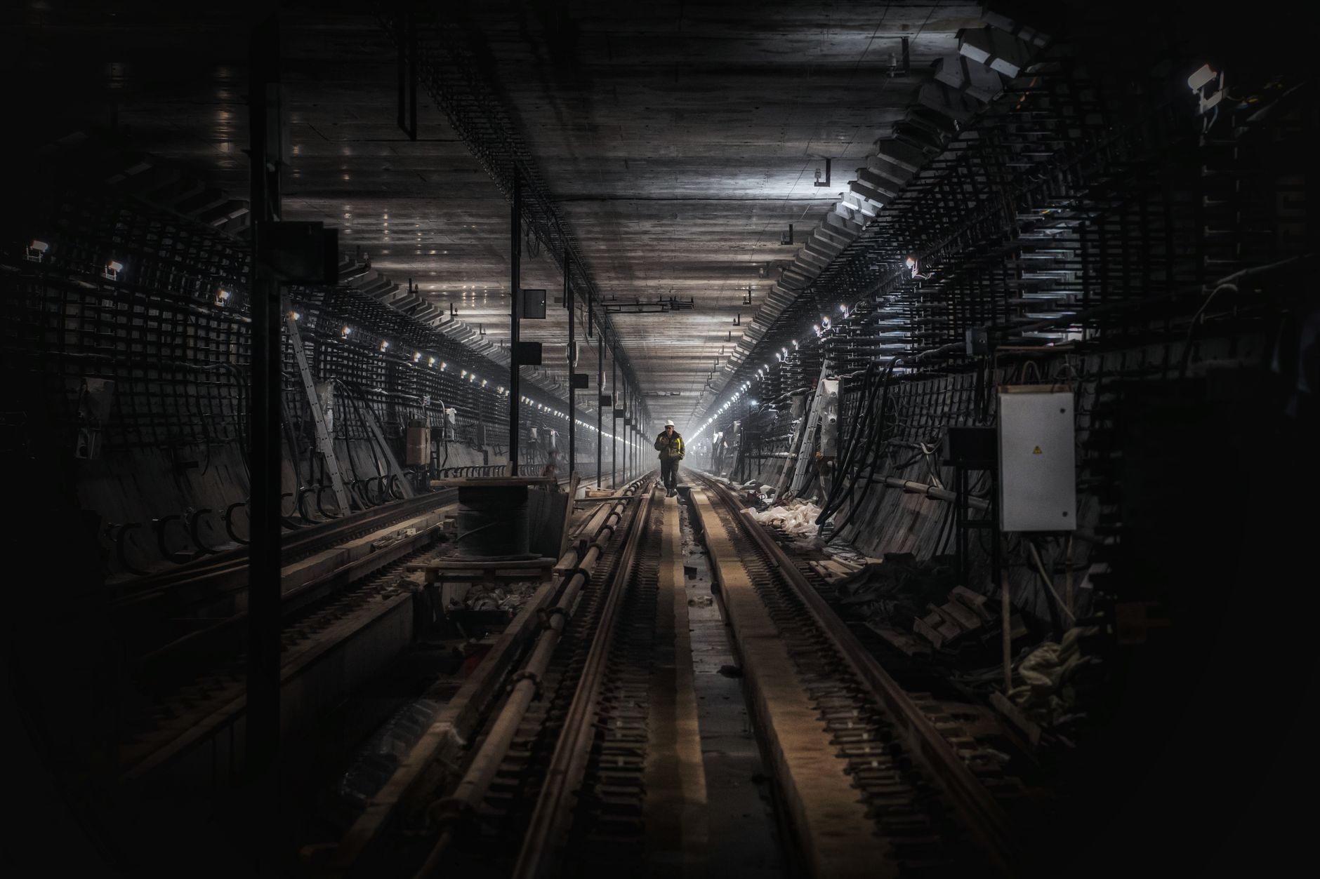 A lone worker walks down train tracks in a dimly lit underground tunnel.
