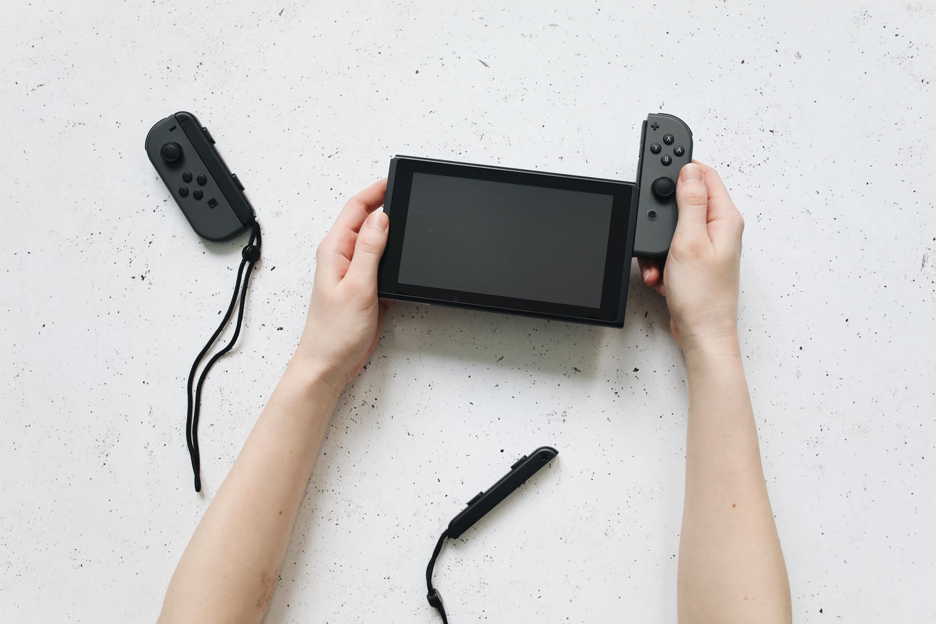 Close-up of hands assembling a Nintendo Switch console with detached controllers on a white surface.
