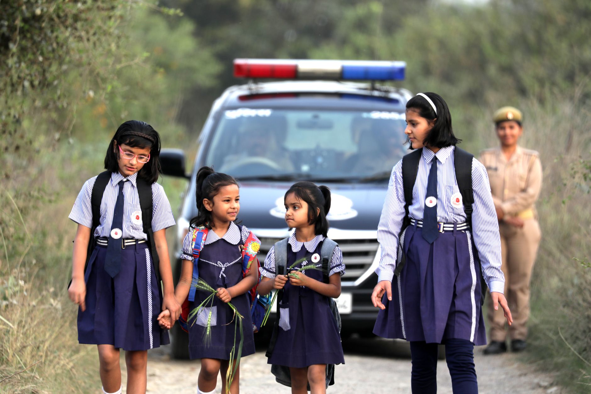 Schoolgirls in uniforms walk safely alongside a police escort on a rural road.