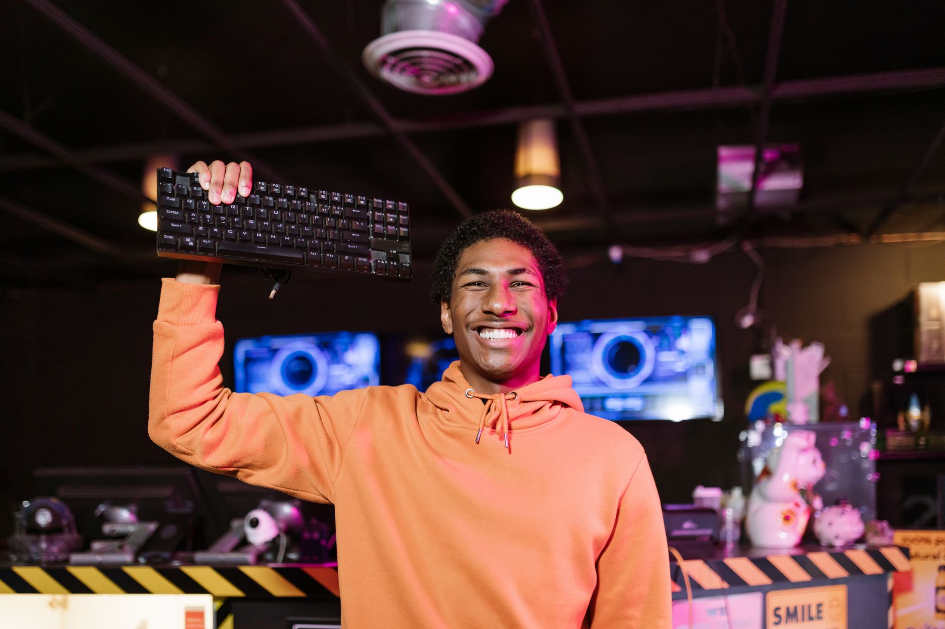 Confident young gamer in orange hoodie holding a keyboard, smiling in a modern esports arena.